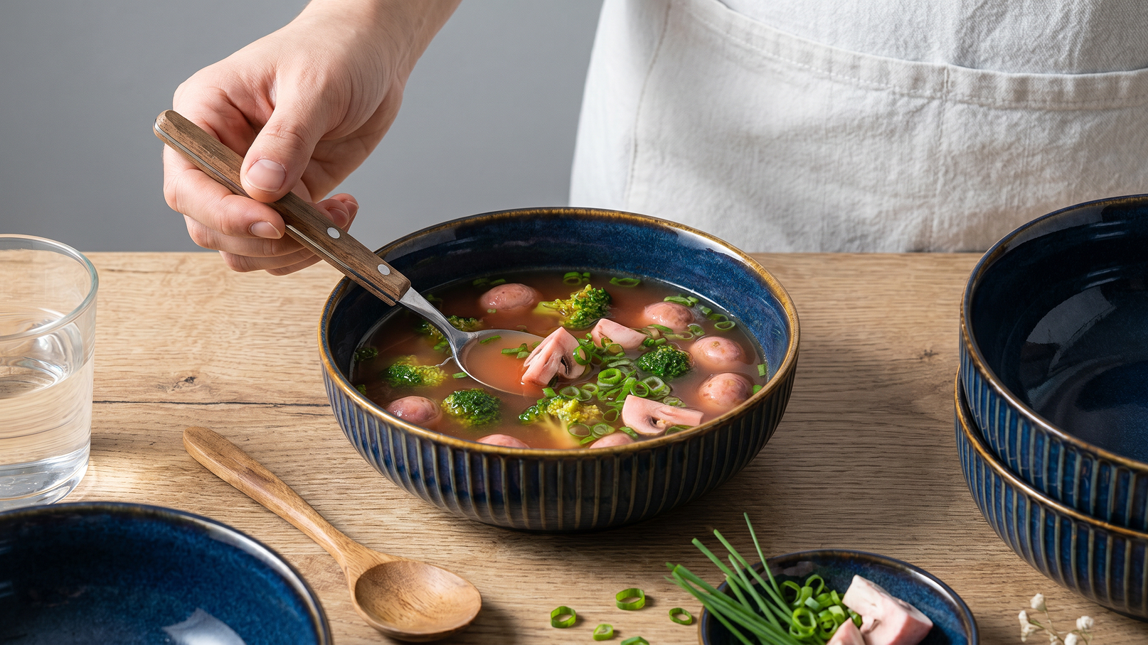 Eine Hand hält einen Löffel und schöpft aus einer tiefen Schüssel mit Brühe. Die Suppe enthält kleine Würstchen, Brokkoli und gehackte Frühlingszwiebeln. Im Hintergrund sind weitere Schalen und ein Glas Wasser zu sehen.