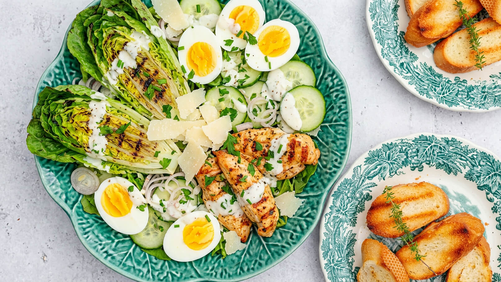 Ein grüner Teller mit gegrilltem Hähnchen, romanesco-Salat, Gurkenscheiben und hartgekochten Eiern. Über dem Salat sind Kräuter und Parmesanraspel verteilt. Neben dem Teller befinden sich zwei kleine Teller mit geröstetem Brot.