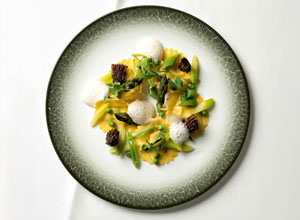 Auf dem Bild ist ein Teller mit frischen Ravioli zu sehen, garniert mit grünem Spargel, Morcheln und kleinen Schaumkugeln. Die Pastagerichte sind in einer ansprechenden Anordnung präsentiert und weisen eine leuchtend gelbe Farbe auf, eingerahmt von einem eleganten Rand.