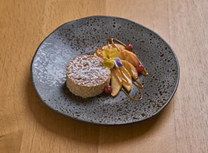 Auf dem Bild ist ein Dessert auf einem dunklen Teller zu sehen. Es besteht aus einem runden, mit Puderzucker bestäubten Kuchen, umgeben von frischen Früchten wie Apfel und dekorativen Blüten. Es sind auch einige Farbtupfer von einer Sauce sichtbar.