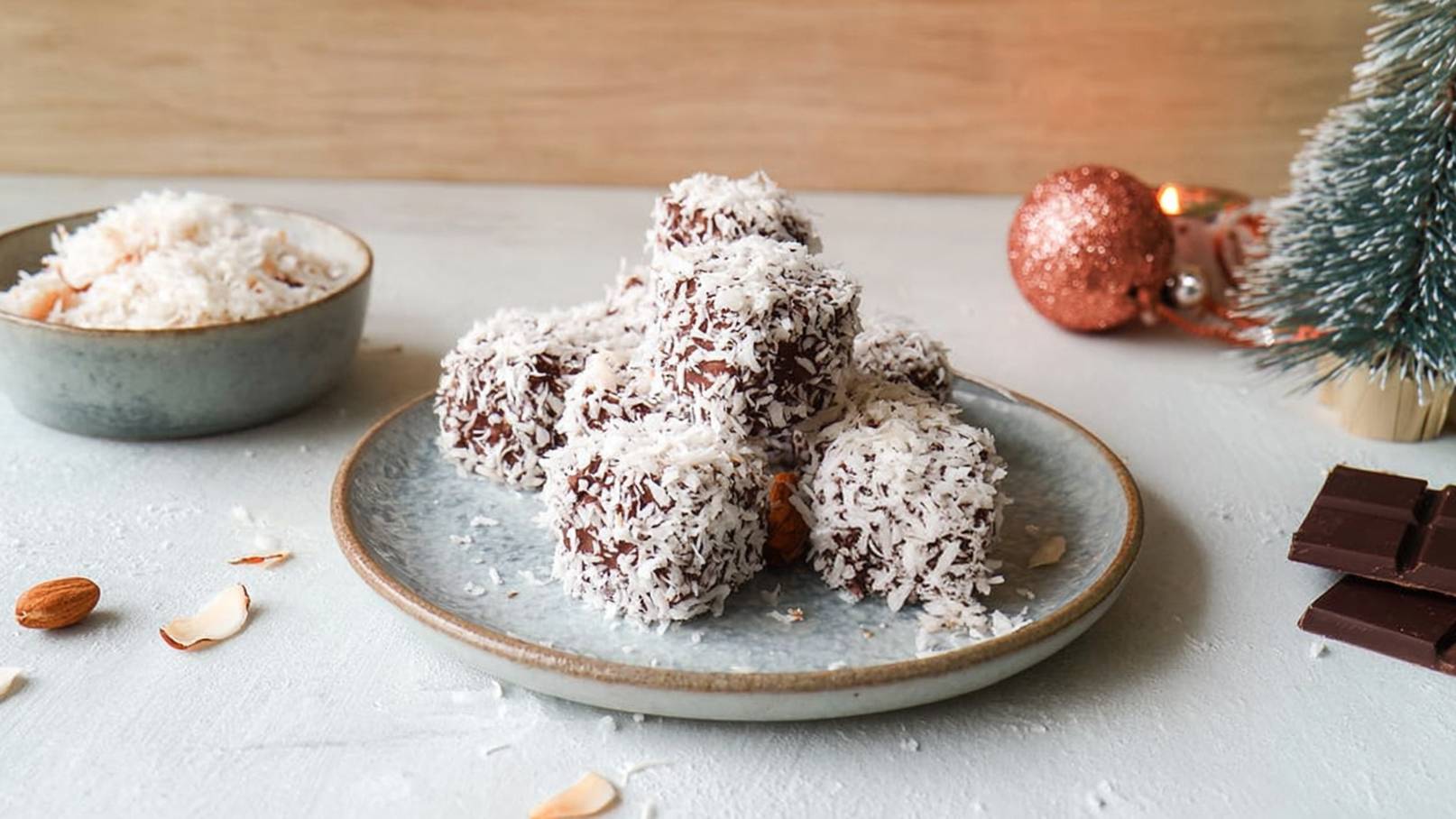Auf einem grauen Teller liegen mehrere quadratische Pralinen, überzogen mit Kokosraspeln. Daneben befindet sich eine Schüssel mit weiteren Kokosraspeln. Die Umgebung ist minimalistisch und weihnachtlich dekoriert.