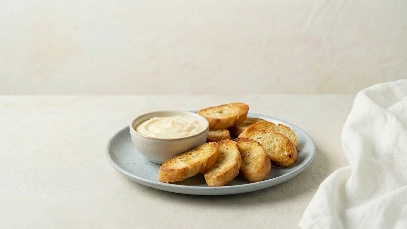 Ein Teller mit gerösteten Brotscheiben liegt neben einer kleinen Schüssel mit einer hellen Dipsauce. Die Brotscheiben sind goldbraun und knusprig. Der Hintergrund ist neutral und lässt die Speisen gut zur Geltung kommen.