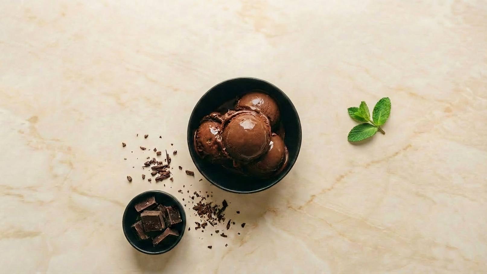 Eine schwarze Schüssel enthält mehrere Kugeln Schokoladeneis. Daneben liegt eine kleinere Schüssel mit Schokoladenstückchen und etwas Schokoladenraspel auf der Tischoberfläche. Ein Blatt frische Minze ergänzt das Bild.