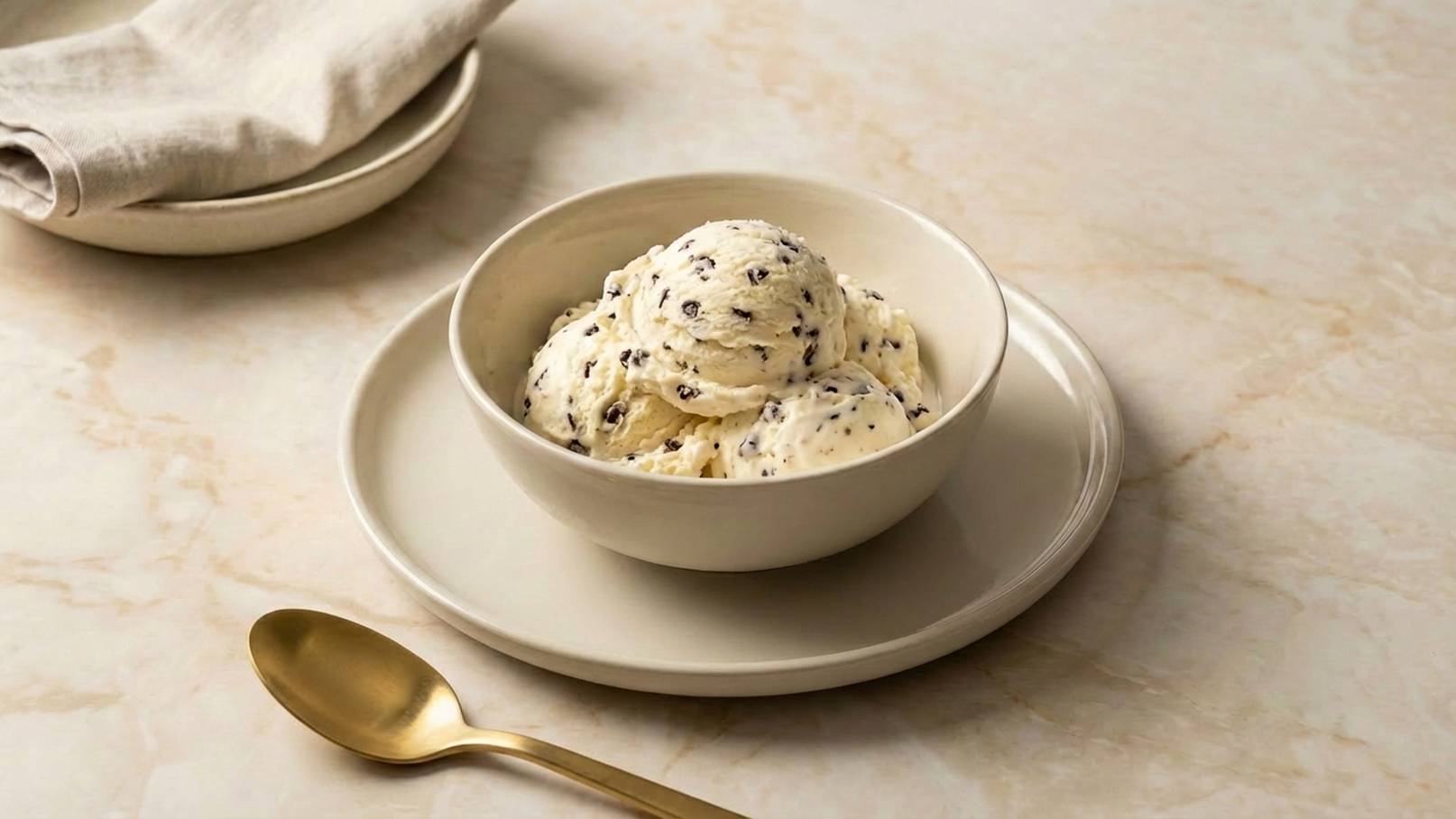 In einer hellen Schüssel auf einem Teller befinden sich drei Kugeln Vanilleeis mit Schokoladenstückchen. Neben der Schüssel liegt ein goldfarbener Löffel. Im Hintergrund liegt ein gedecktes Tuch und ein weiterer Teller.