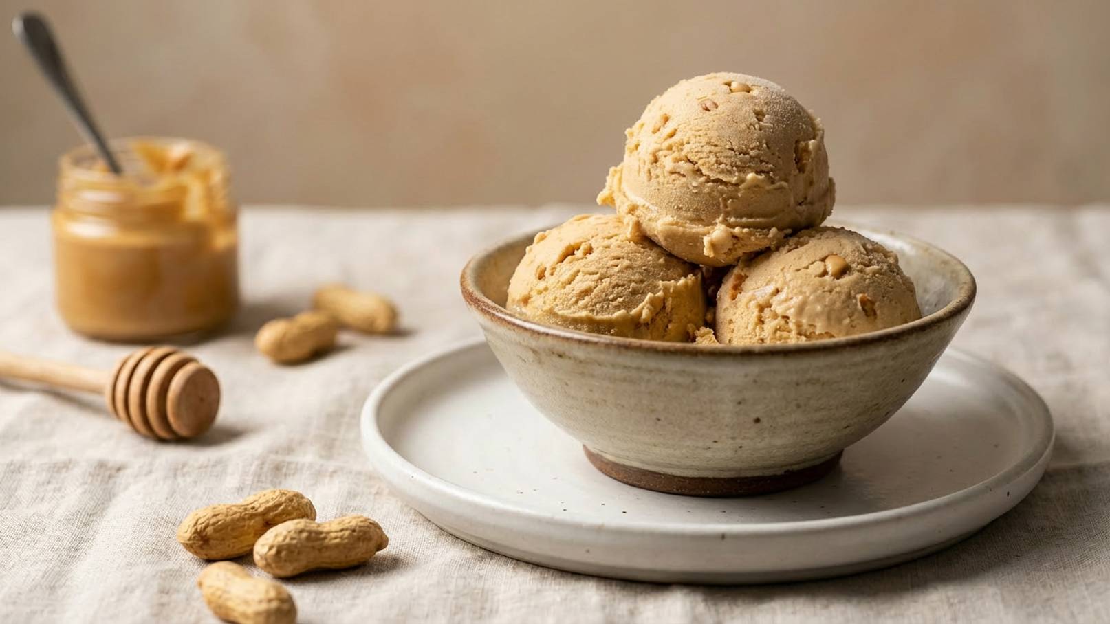 Eine Schüssel mit drei Kugeln Erdnusseis steht auf einem Teller. Daneben befindet sich ein Glas mit Erdnussbutter und ein Honiglöffel. Um die Schüssel liegen einige Erdnüsse. Der Hintergrund ist weich und neutral gehalten.