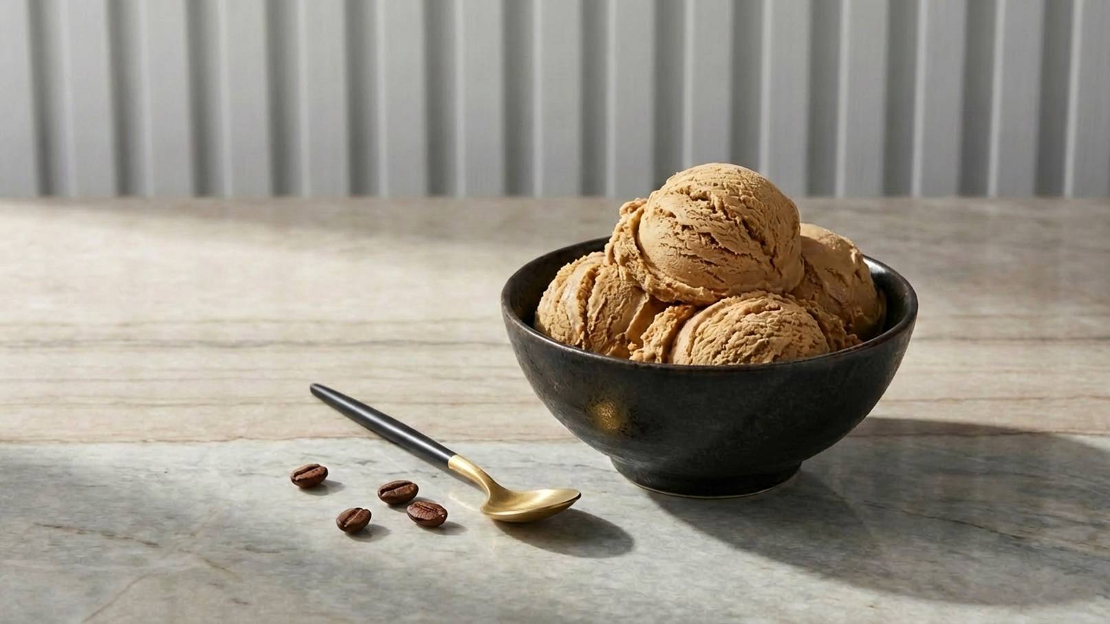 Eine schwarze Schale mit mehreren Kugeln Kaffeeeis steht auf einem Tisch. Neben der Schale liegen einige Kaffeebohnen und ein goldener Löffel. Der Hintergrund zeigt eine helle, vertikale Struktur.