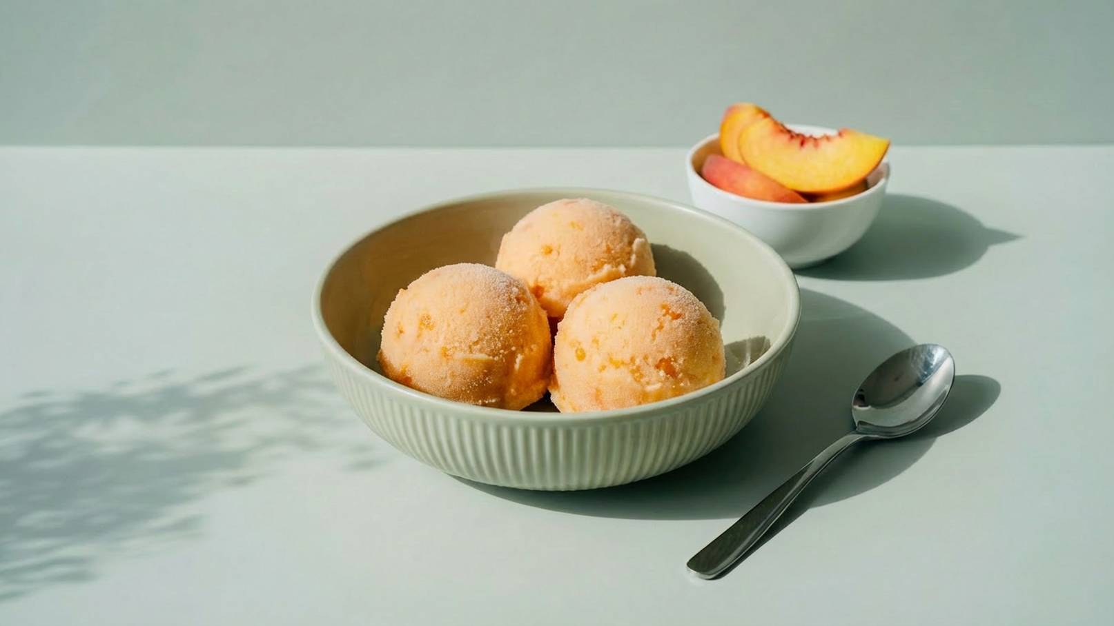 Eine Schüssel mit drei Farben von Pfirsich-Eis auf einem hellen Tisch. Daneben steht eine kleine Schale mit frischen Pfirsichscheiben und ein Löffel liegt bereit.