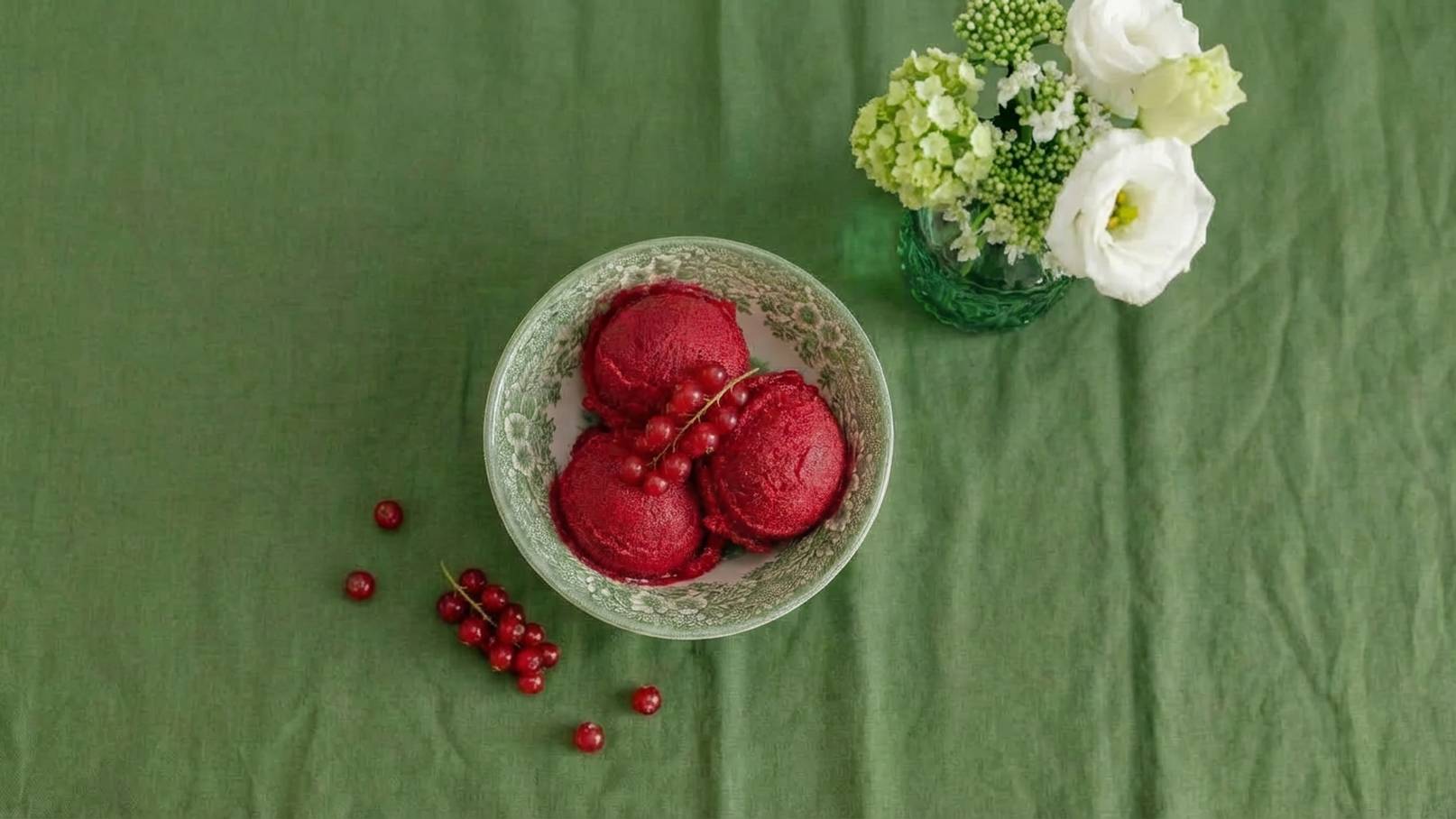 Eine grüne Tischdecke zeigt eine dekorative Schale mit drei Kugeln rotem Sorbet, verziert mit frischen roten Johannisbeeren. Neben der Schale liegt ein kleiner Haufen von Johannisbeeren. Im Hintergrund steht ein Glas mit weißen und grünen Blumen.