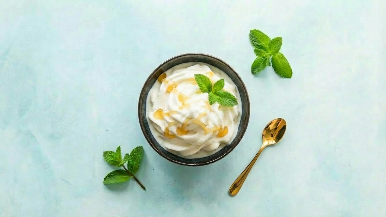 Eine schwarz-braune Schüssel enthält cremigen Joghurt, garniert mit frischer Minze und einem Tropfen Honig. Daneben liegt ein kleiner goldfarbener Löffel und einige Minzblätter sind um die Schüssel verteilt. Der Hintergrund ist hellblau.