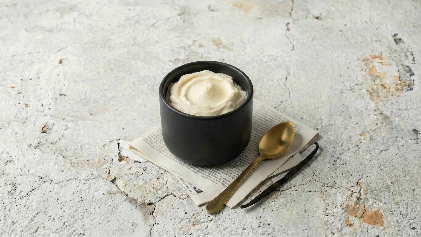 Ein schwarzer Schüssel mit cremigem, hellem Inhalt steht auf einem Stapel Zeitung. Daneben liegt ein goldener Löffel. Der Hintergrund ist unregelmäßig und strukturiert, was die Schüssel hervorhebt.