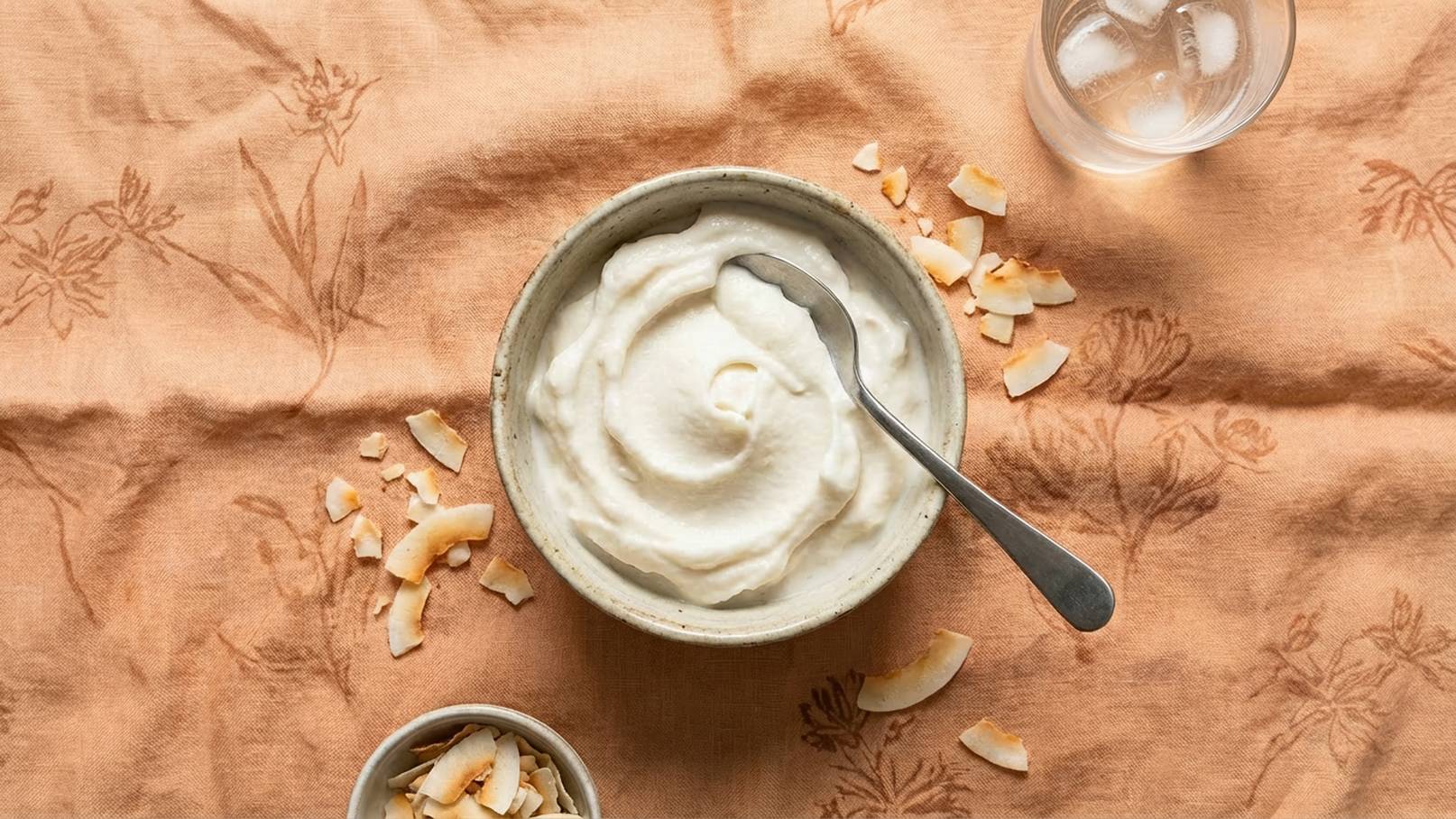 Eine Schüssel mit cremigem Joghurt steht auf einem orangefarbenen Tuch. Neben der Schüssel liegen gehobelte Kokosnüsse. Ein Löffel steckt im Joghurt. Daneben befindet sich ein Glas mit Eiswasser.