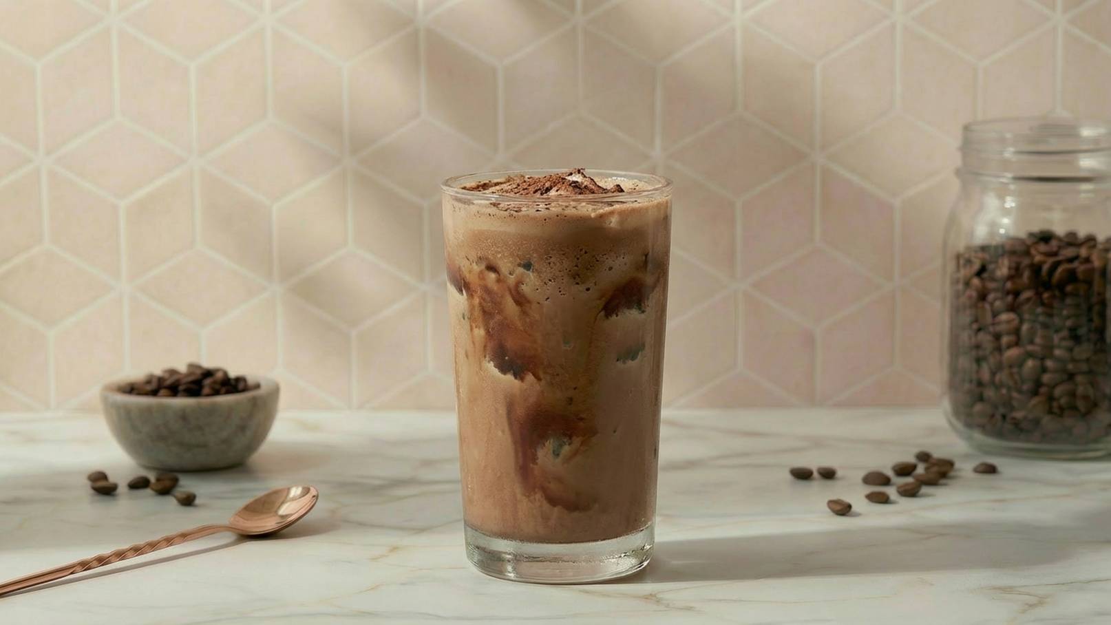 Ein Glas kalter Kaffee mit Schaum und Schokoladenstückchen steht auf einem Tisch. Im Hintergrund sind Kaffeebohnen in einer Schale und ein Glasbehälter sichtbar. Die Wand hinter dem Glas hat ein geometrisches Muster.
