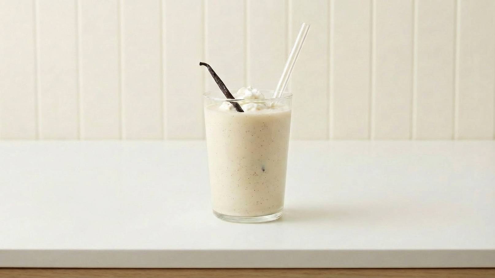 Ein Glas mit einem cremigen Getränk, garniert mit Schlagsahne und einer Vanilleschote. Zwei Strohhalme stecken im Glas. Der Hintergrund ist hell und neutral, was den Fokus auf das Getränk lenkt.