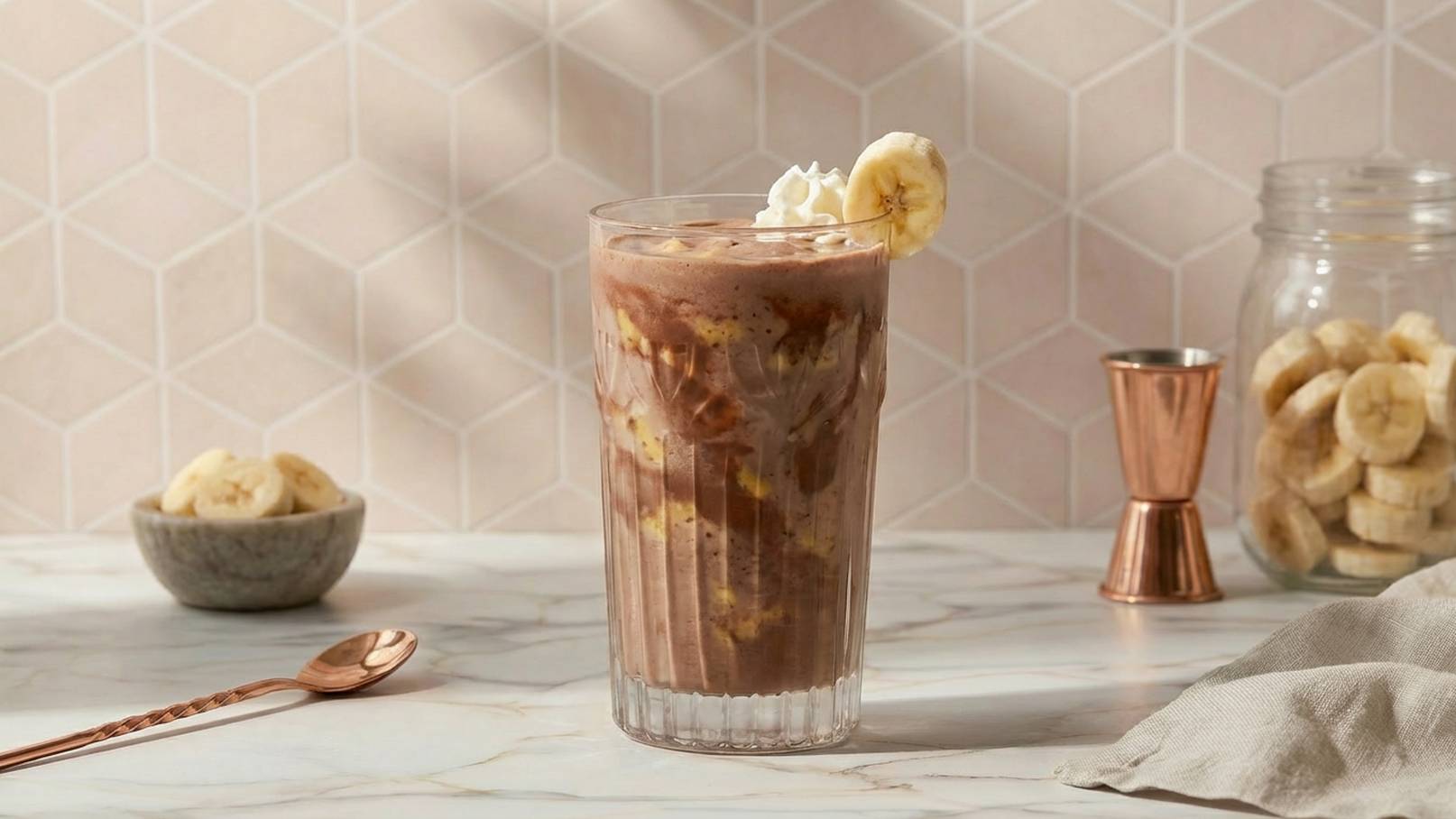 Ein Glas mit einem schokoladigen Bananenshake, garniert mit Sahne und einer Bananenscheibe. Im Hintergrund befindet sich eine Schüssel mit getrockneten Bananen und ein Glasbehälter mit weiteren Bananenscheiben. Die Oberfläche ist marmoriert, und die Wand hat ein geometrisches Muster.