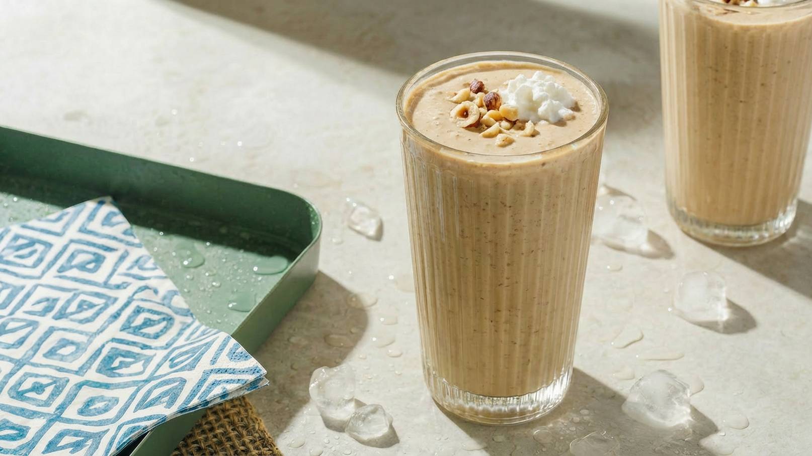 Zwei große Gläser mit einem cremigen, nussigen Getränk stehen auf einem Tisch. Eines der Gläser ist mit Sahne und Nüssen garniert. Daneben liegt eine bunte Serviette und ein Tablett. Wasserflecken deuten auf Frische hin.