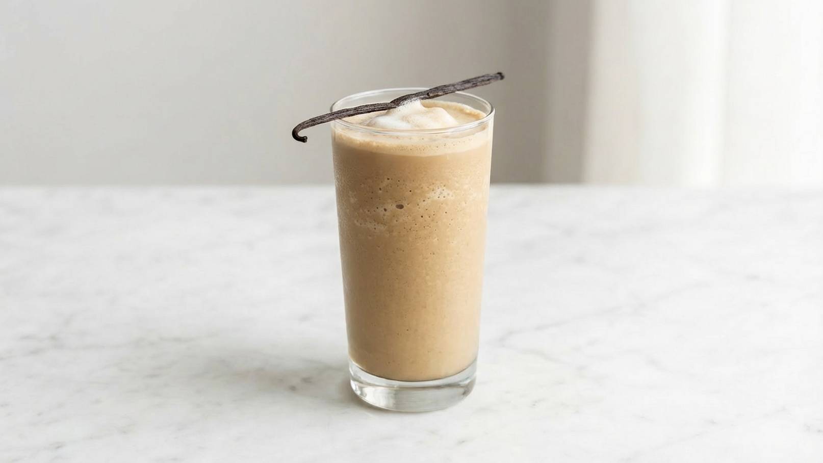 Ein Glas mit einem cremigen, braunen Getränk, das eine Schaummütze hat. Eine Vanilleschote liegt dekorativ auf dem Rand des Glases. Der Hintergrund ist neutral und konzentriert sich auf das Getränk.