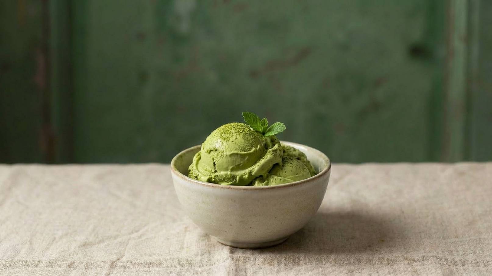Eine Schale mit grünem Eis, garniert mit einem frisch gebetteten Minzblatt, steht auf einem beigefarbenen Tisch. Der Hintergrund ist unscharf und leicht grün. Das Eis hat eine cremige Textur und sieht appetitlich aus.