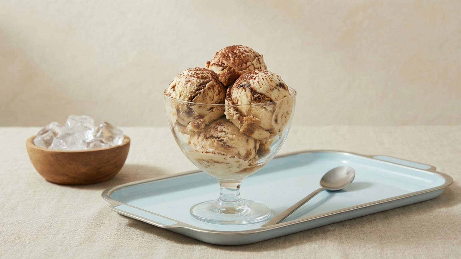 Eine Schüssel mit mehreren Kugeln Schokoladeneis, garniert mit Kakaopulver, steht auf einem blauen Tablett. Daneben befindet sich eine Schüssel mit Eiswürfeln und ein Löffel. Der Hintergrund ist neutral und weich.