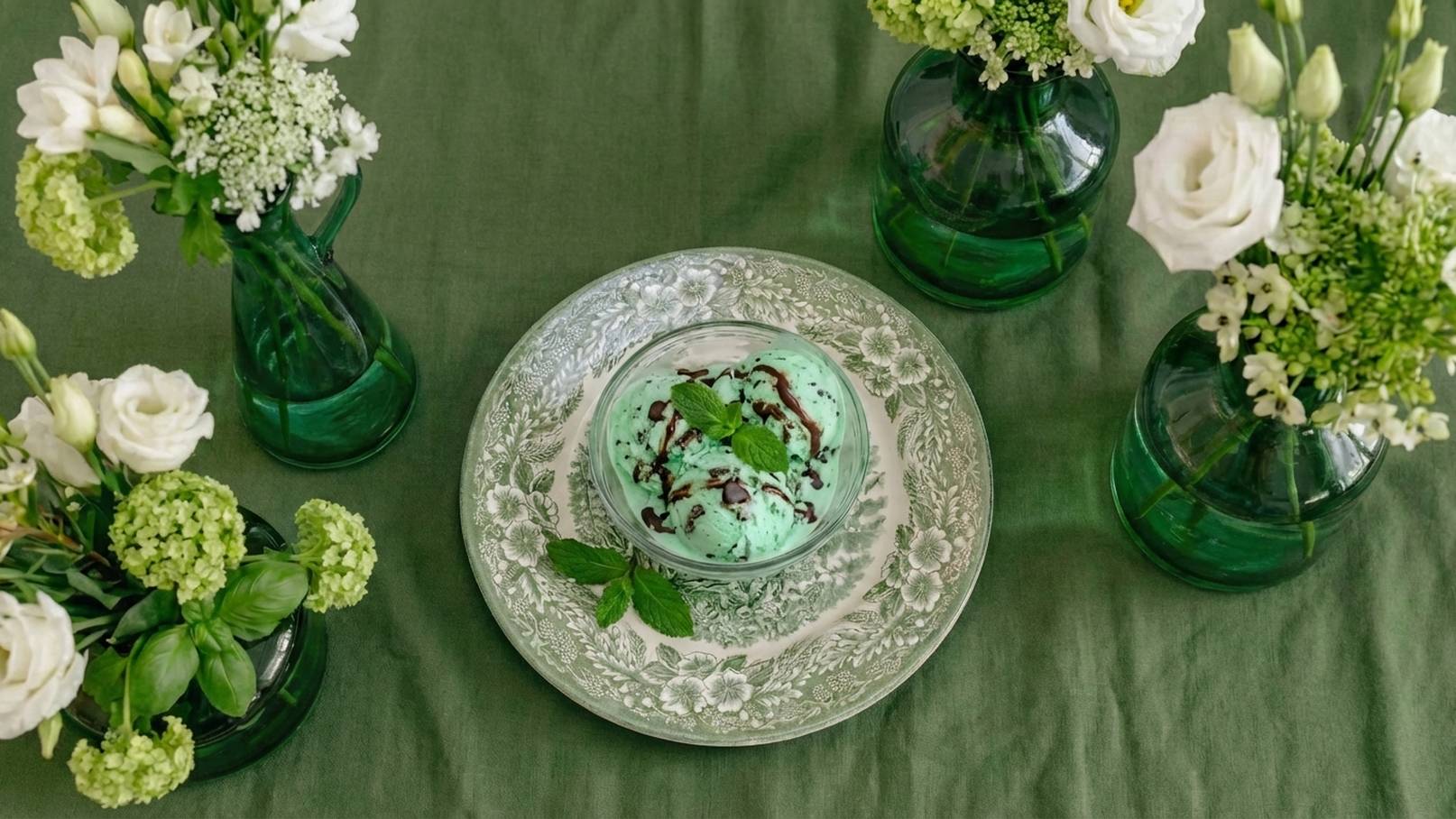 Eine grüne Tischdecke umgibt einen silbernen Teller mit einer Portion mintgrünem Eis, garniert mit Schokolade und frischer Minze. Um den Teller herum stehen mehrere grüne Glasvasen mit weißen Blumen und grünen Pflanzen, die eine frische und einladende Atmosphäre schaffen.