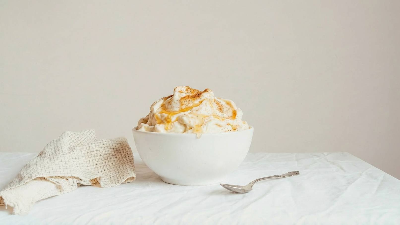 Eine große Schüssel mit cremigem Dessert, garniert mit einer goldenen Karamellsoße, steht auf einem weißen Tisch. Neben der Schüssel liegt ein Löffel und ein Küchenhandtuch. Der Hintergrund ist schlicht und neutral gehalten.