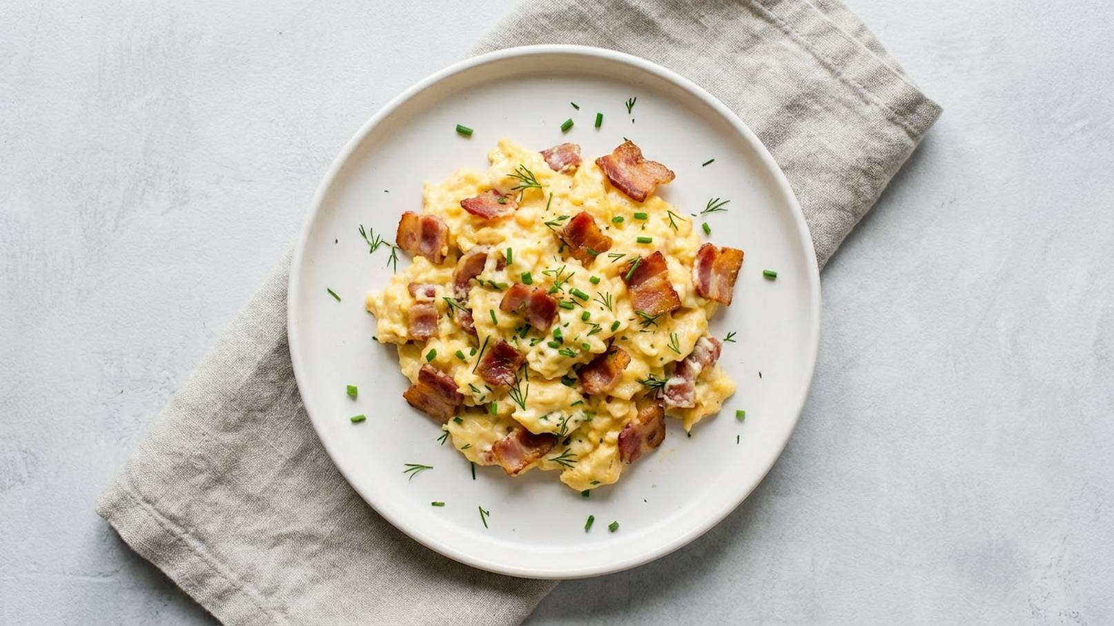 Ein Teller mit cremigen Rühreiern, garniert mit knusprigem Speck und frischem Schnittlauch. Die Eier erscheinen goldgelb und sind gut vermischt, während die Speckstücke eine knusprige Textur verleihen. Der Teller steht auf einem neutralen Untergrund.