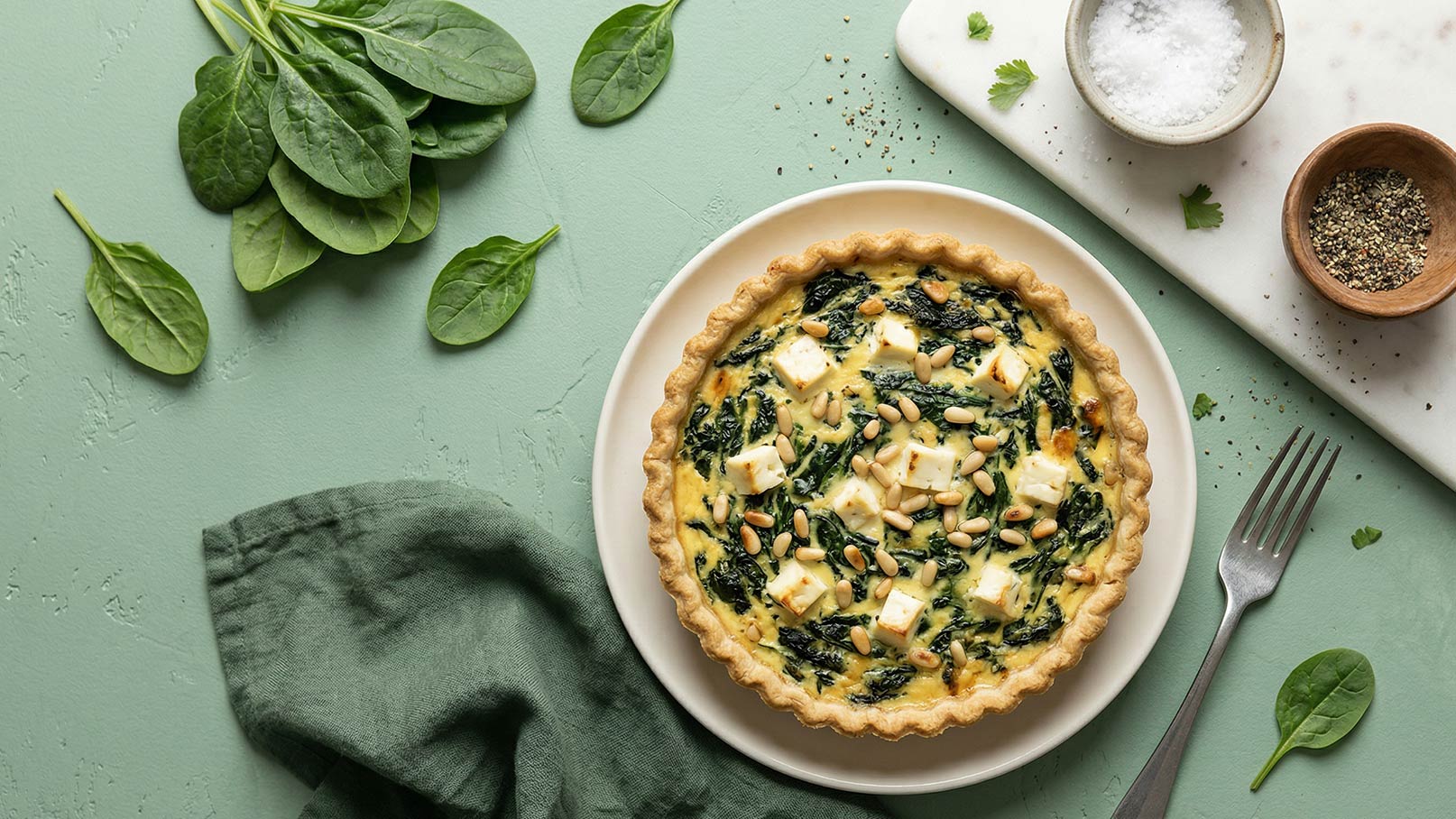 Ein herzhaftes Spinatquiche auf einem weißen Teller. Die Quiche ist gefüllt mit Spinat, Feta und Pinienkernen. Daneben liegen frische Spinatblätter und Gewürze auf einem Holzbrett. Eine grüne Serviette und eine Gabel sind ebenfalls sichtbar.