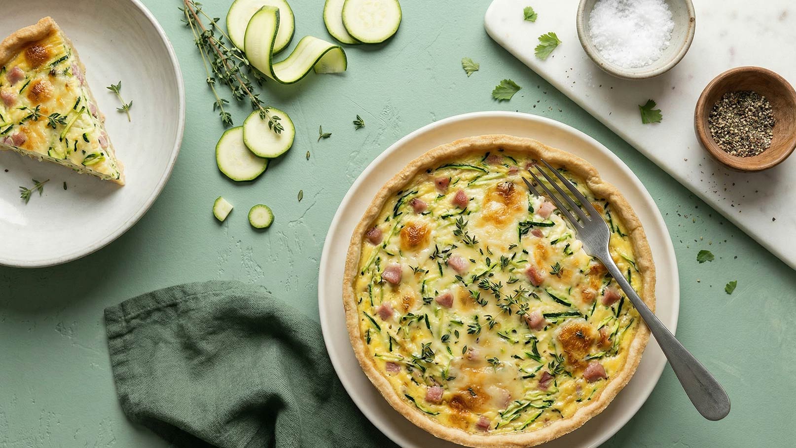 Ein Teller mit einer frisch gebackenen Quiche, gefüllt mit Zucchini, Schinken und Kräutern. Neben der Quiche liegt ein Stück auf einem separaten Teller. Farblich harmonische grüne Unterlage und einige Zucchinischeiben auf dem Tisch ergänzen das Bild.