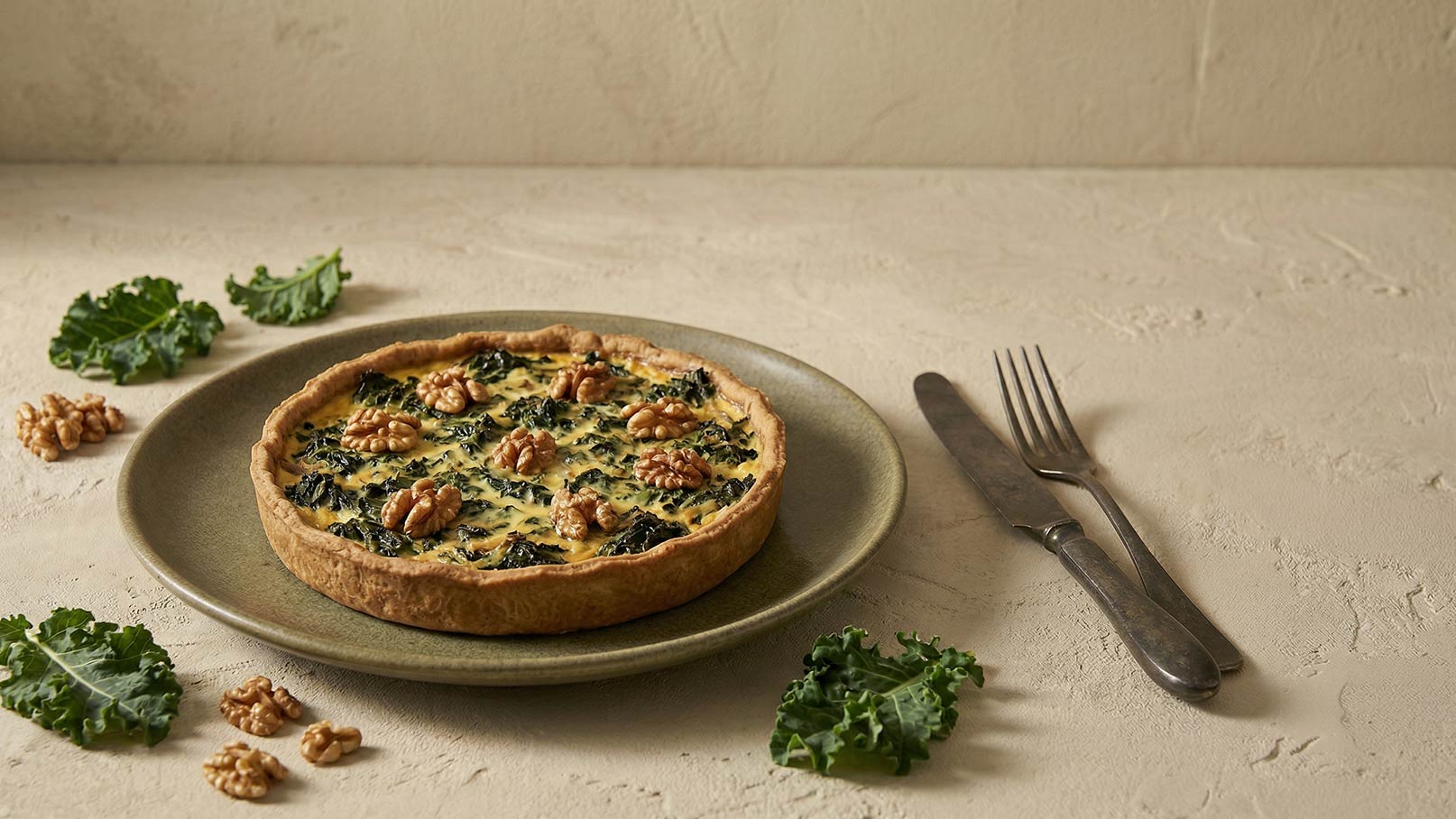 Eine herzhafte Quiche mit einem Mürbeteigboden, gefüllt mit grünem Blattgemüse und Walnüssen. Sie liegt auf einem flachen Teller. Einige frische Grünkohlblätter und Walnüsse sind um den Teller angeordnet, und Besteck liegt daneben.