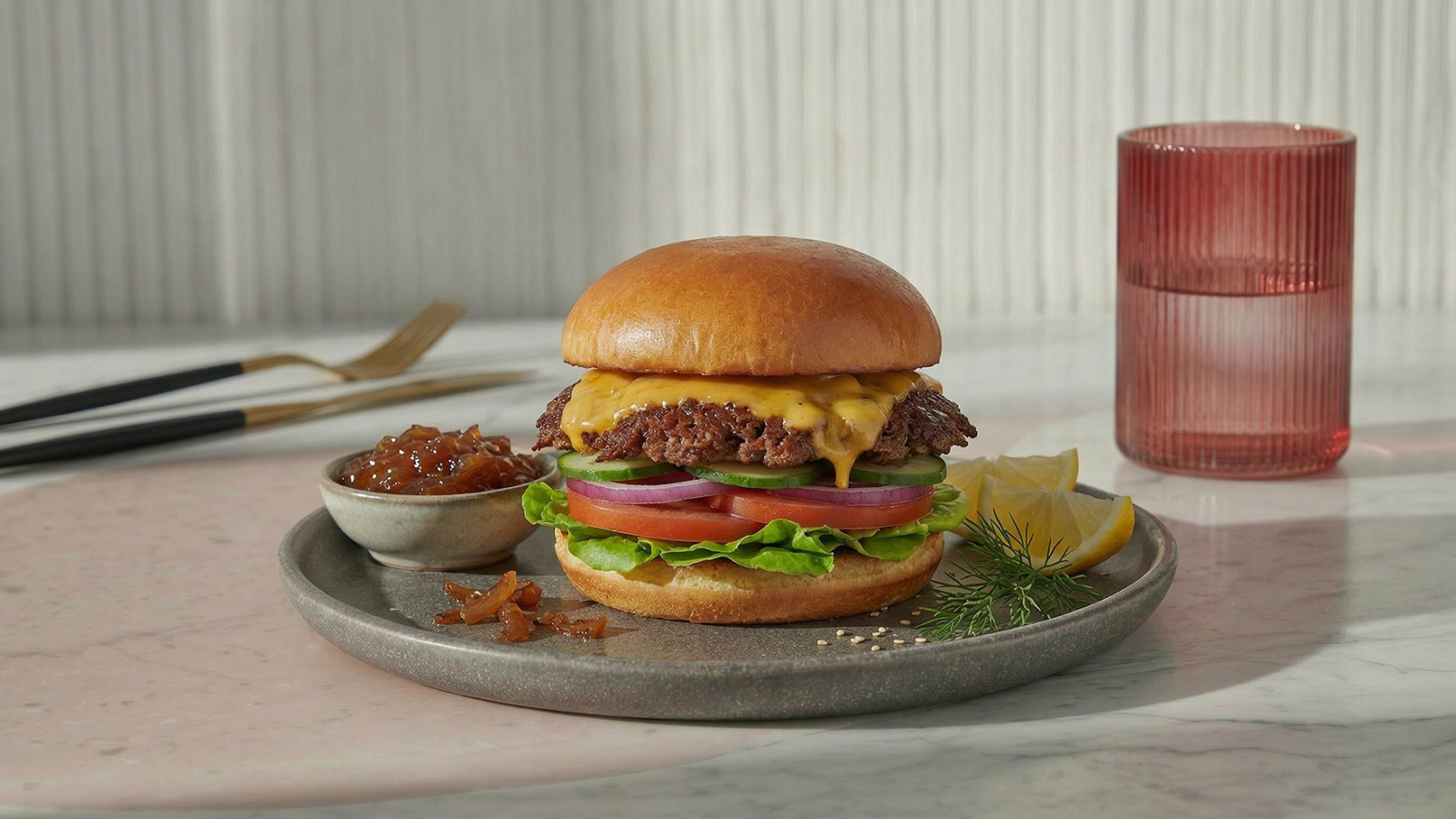 Ein saftiger Hamburger mit einer Käsescheibe, frischem Gemüse wie Salat und Zwiebeln, serviert auf einem grauen Teller. Daneben befindet sich eine kleine Schale mit einer süßen Sauce und eine Zitronenscheibe. Ein Glas Wasser steht im Hintergrund.