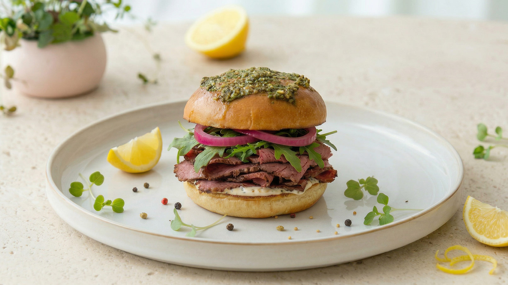 Ein saftiger Burger auf einem weißen Teller, gefüllt mit roastbeef, roten Zwiebeln und Rucola. Oben ist eine grüne Kräuterpaste. Neben dem Teller liegen Zitronenscheiben und kleine Pflanzen. Der Hintergrund ist hell und weich, was eine einladende Atmosphäre schafft.