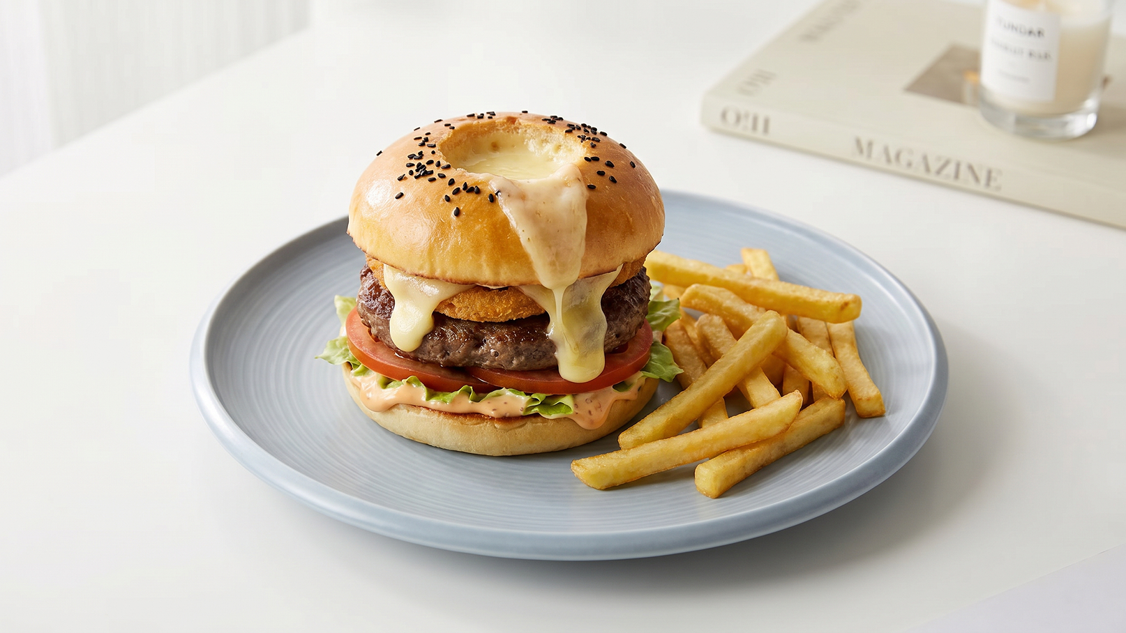 Auf einem blauen Teller liegt ein großer Burger mit Käse, einer Fleischscheibe, Tomate und Salat. Daneben befinden sich frittierte Pommes. Der Burger hat ein weiches Brötchen mit Sesam.