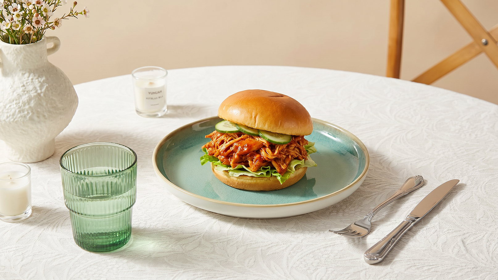 Ein gefülltes Burger-Brötchen mit zerrissenem Fleisch, Salat und Gurkenscheiben liegt auf einem blauen Teller. Der Tisch ist mit einer weißen Tischdecke gedeckt, und daneben stehen ein grünes Glas und ein kleines weißes Kerzenlicht.