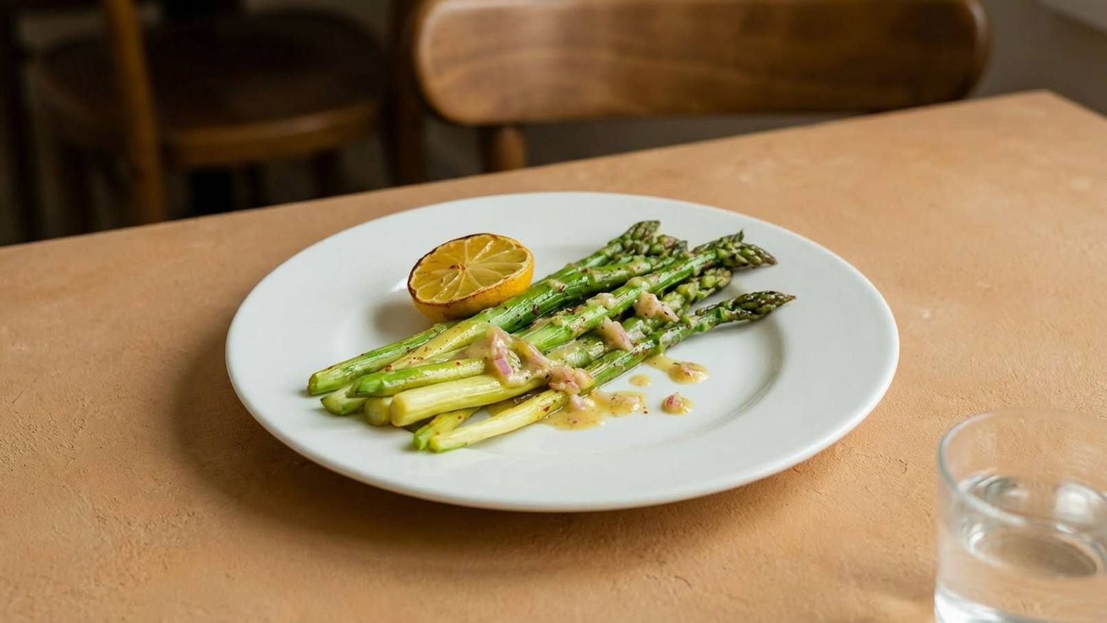 Ein Teller mit grünem Spargel, der mit einer Sauce überzogen ist. Eine halbe Zitrone liegt neben dem Spargel. Der Teller steht auf einem hellen Tisch.