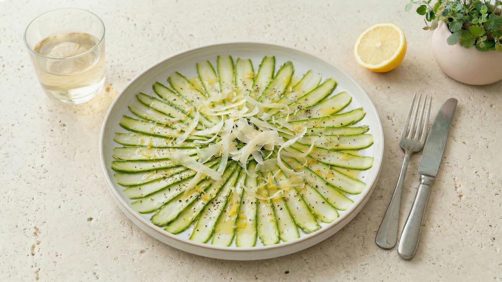 Ein Teller mit grünem Spargel, der mit einer hellen Sauce beträufelt ist. Der Teller befindet sich auf einer marmorierten Oberfläche. Daneben steht ein Glas Wasser. Der Hintergrund ist aus Holz.