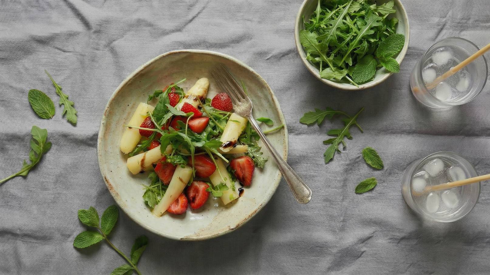Eine Schüssel mit frischem Salat, bestehend aus Spargel, Erdbeeren und Rucola, steht im Vordergrund. Daneben befindet sich eine weitere Schüssel mit grünem Salat. Im Hintergrund sind zwei Gläser mit Eiswasser und Strohhalmen zu sehen.
