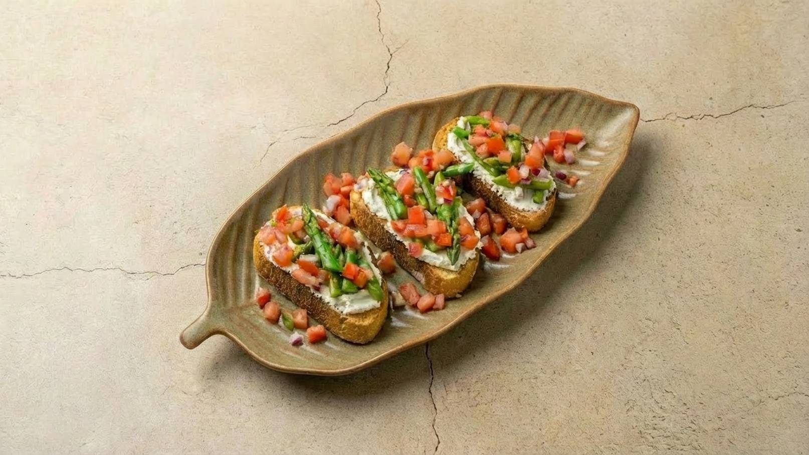 Drei belegte Brote auf einer länglichen, blattförmigen Platte. Die Brote sind mit frischen Tomatenstücken, Kräutern und einer cremigen Sauce garniert, was ein appetitliches Gericht darstellt. Der Untergrund ist neutral und hebt die Farben der Zutaten hervor.