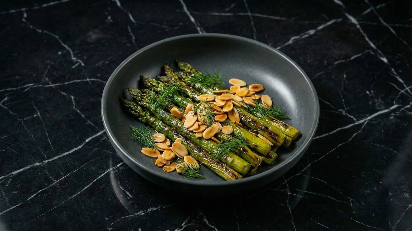 Ein schwarzes Teller mit gegrilltem Spargel, garniert mit frischem Dill und gerösteten Mandelblättchen. Der Spargel ist goldbraun und knackig, während die Mandeln für einen zusätzlichen Crunch sorgen. Der Kontrast zwischen dem Gemüse und dem Teller hebt die Farben hervor.