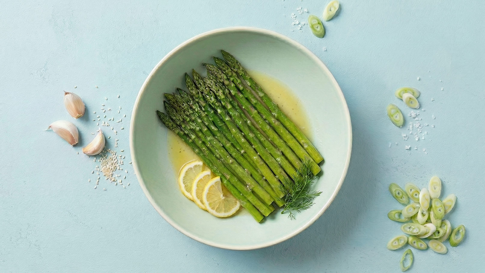 Ein weißer Spargel auf einem weißen Teller, serviert mit einer Soße und garniert mit gehacktem Schnittlauch. Im Hintergrund sind einige Orangenscheiben und eine Schale mit grobem Salz sichtbar.