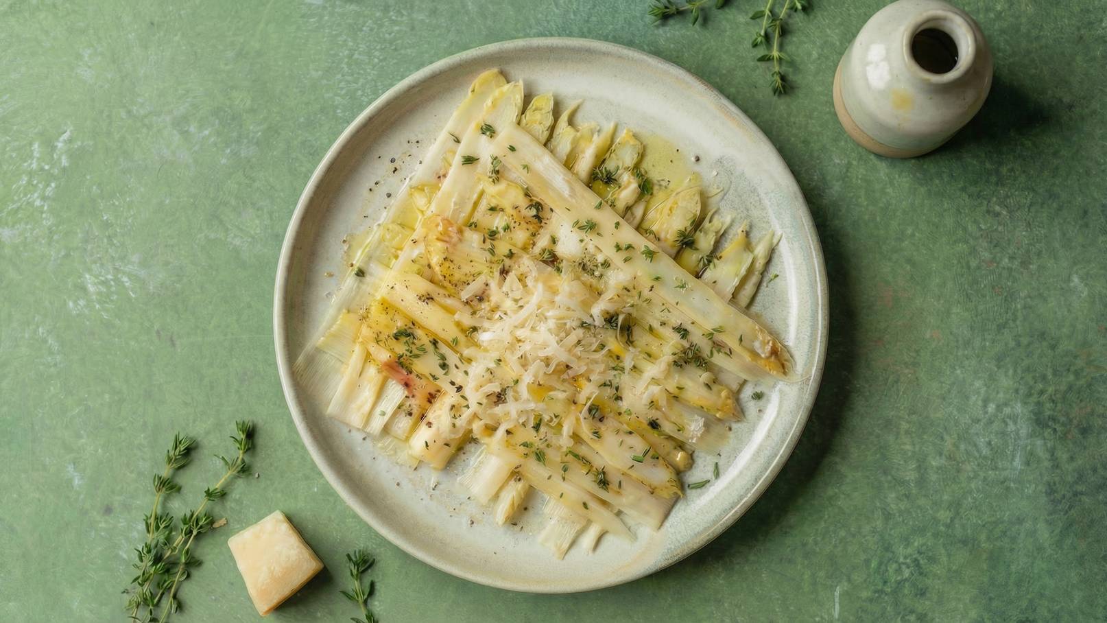 Ein Teller mit Spargel, der mit gehobeltem Käse und Kräutern garniert ist. Der Teller steht auf einer grünen Oberfläche, umgeben von frischen Kräutern und einem kleinen Gefäß, das vielleicht Öl oder Essig enthält.