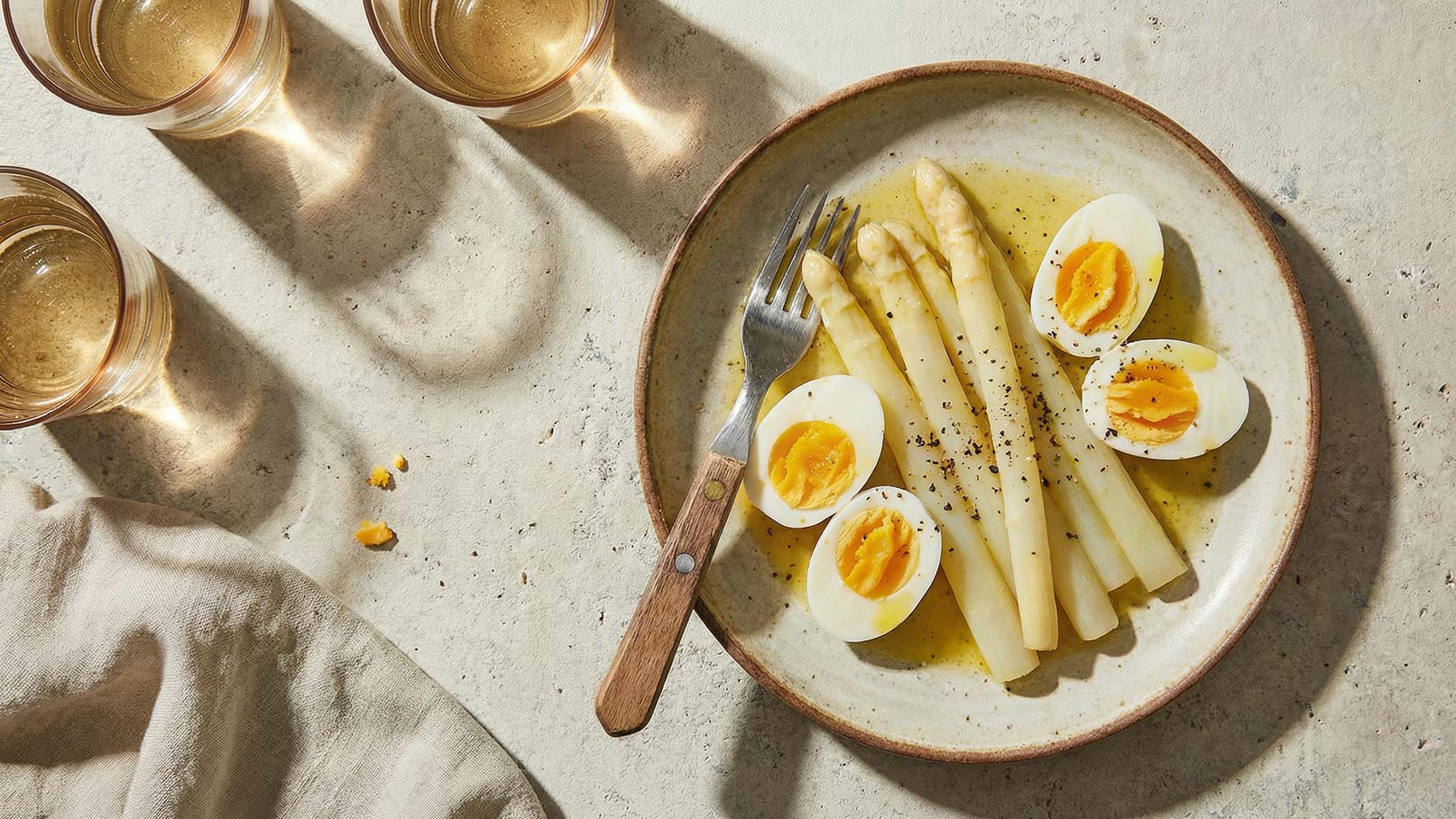 Ein Teller mit weißen Spargelstangen, die zusammen mit halbierten gekochten Eiern serviert sind. Die Eier sind mit einer gelben Eigelbfüllung sichtbar. Im Hintergrund sind Gläser mit Getränk sichtbar. Das Gericht ist mit etwas Pfeffer gewürzt.