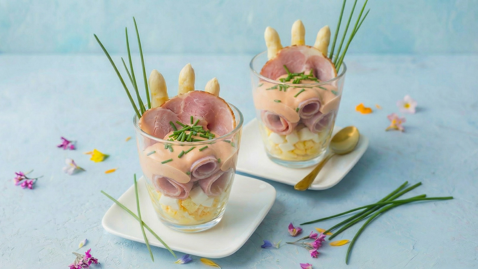 Zwei Gläser mit Schichten aus gekochtem Ei, einer cremigen Füllung und Scheiben von Schinken. Auf jedem Glas befinden sich einige Spargelspitzen und frisch gehackter Schnittlauch. Die Gläser stehen auf weißen Unterteller, umgeben von bunten Essblüten.