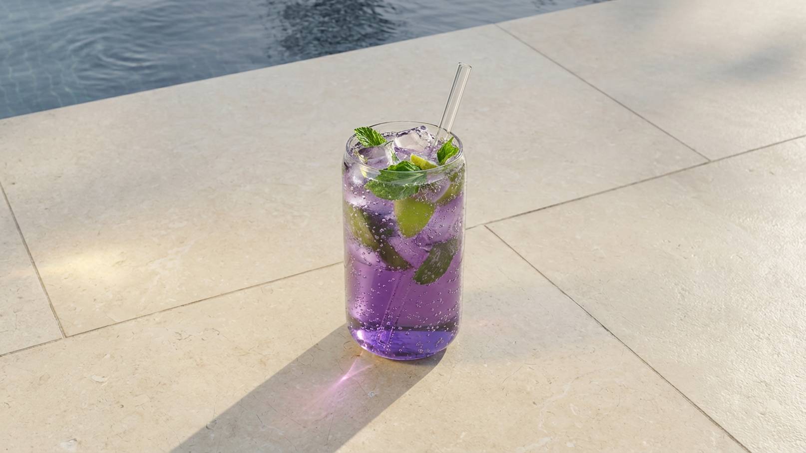 Ein Glas mit lila Getränk steht auf einem hellen Steinuntergrund. Das Getränk enthält frische Minzblätter und Limettenscheiben, mit Sprudelwasser versehen. Im Hintergrund ist ein Schwimmbecken sichtbar.
