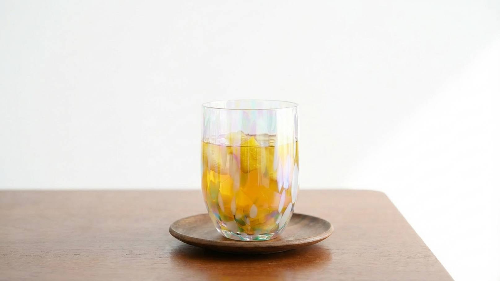 Ein transparentes Trinkglas mit einem gelben Getränk und Eiswürfeln steht auf einem runden Holzuntersetzer. Der Hintergrund ist neutral und hell.