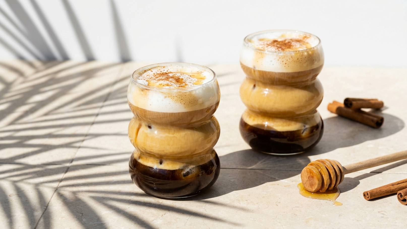 Zwei gläserne Becher mit geschichtetem Kaffee, Sahne und einer Karamellnote stehen auf einem hellen Tisch. Zusätzlich liegen ein Holzlöffel und Zimtstangen neben den Bechern. Die Schatten der Umgebung fallen sanft auf die Fläche.