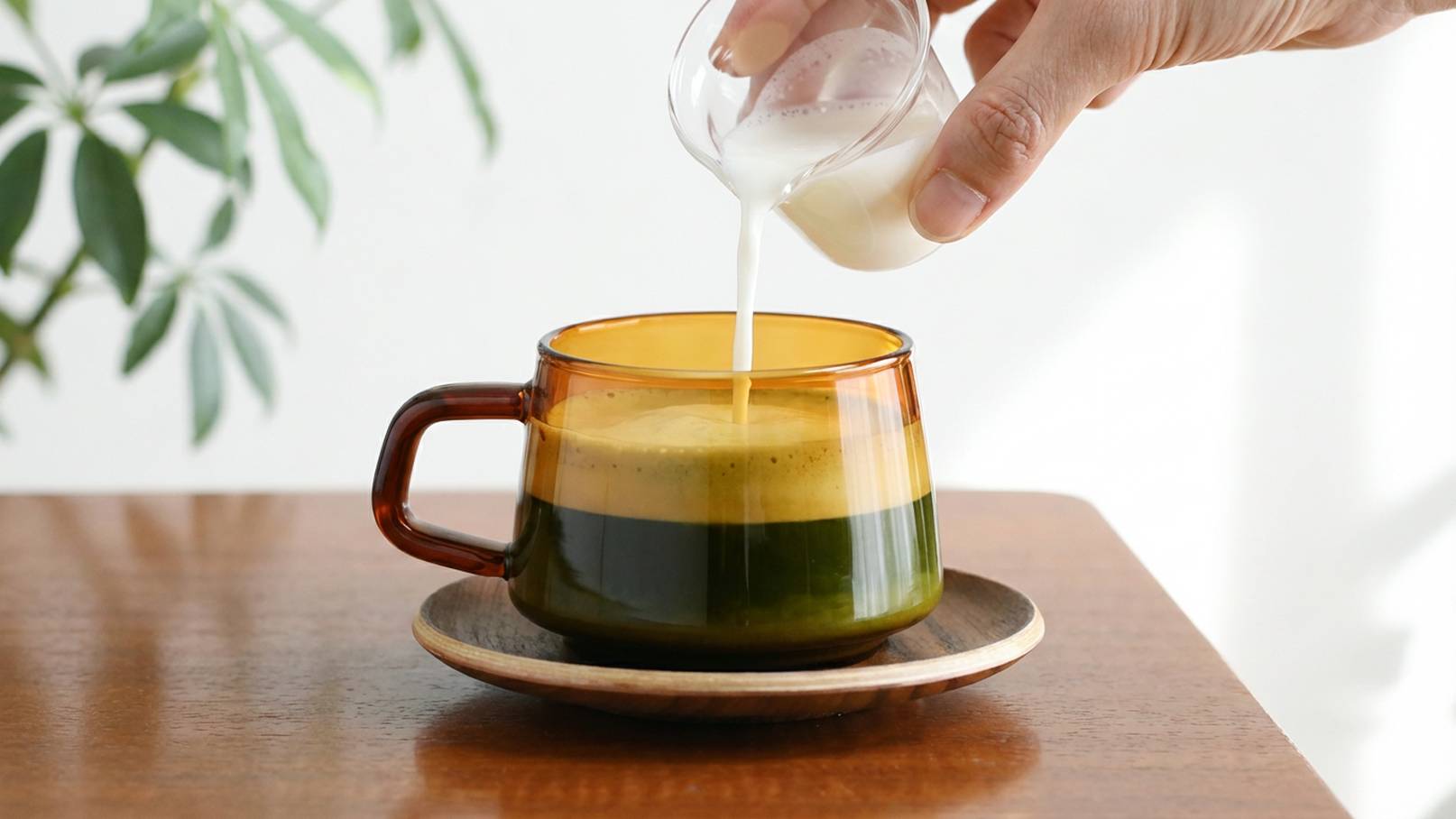 Eine Hand gießt Milch aus einem kleinen Behälter in eine mehrfarbige Tasse mit einer Schicht aus grünem Getränk und einer schaumigen Oberseite. Die Tasse steht auf einem Holzuntersetzer auf einem Tisch. Im Hintergrund ist eine Pflanze sichtbar.