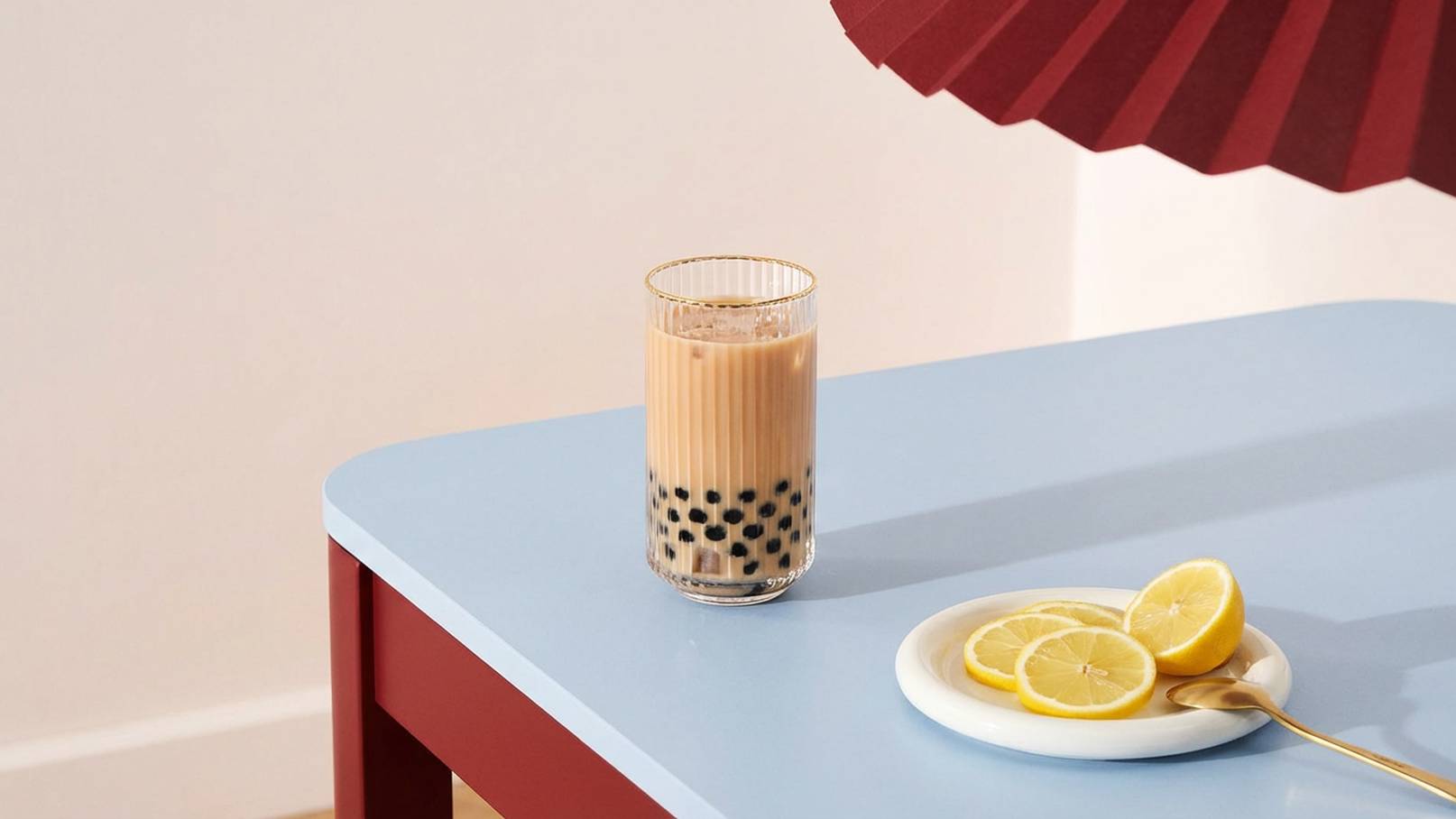 Ein Glas mit Milchtee und Tapiokaperlen steht auf einem blauen Tisch. Neben dem Glas liegt ein Teller mit zwei Scheiben Zitrone und einem kleinen Löffel. Der Hintergrund ist hell und minimalistisch.