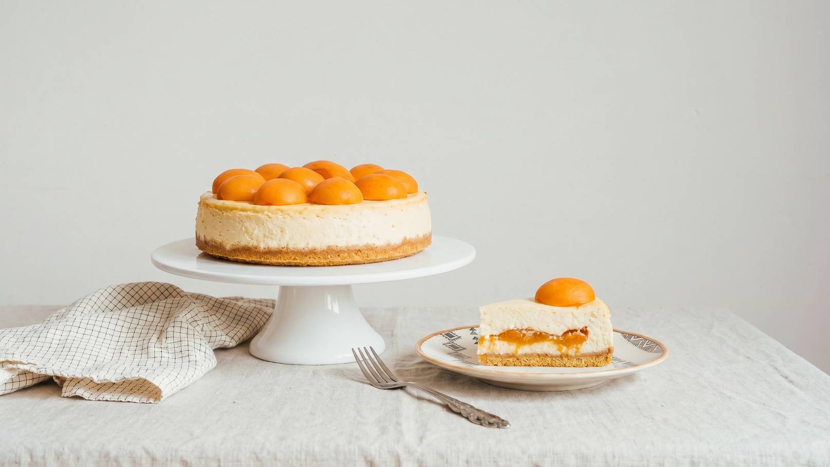 Auf einem weißen Kuchenteller steht ein Käsekuchen, dekoriert mit kleinen, orangenen Früchten. Neben dem Kuchen liegt ein Stück Käsekuchen auf einem Teller, mit einer Frucht obenauf. Ein Besteck liegt daneben auf einer gedeckten Oberfläche.