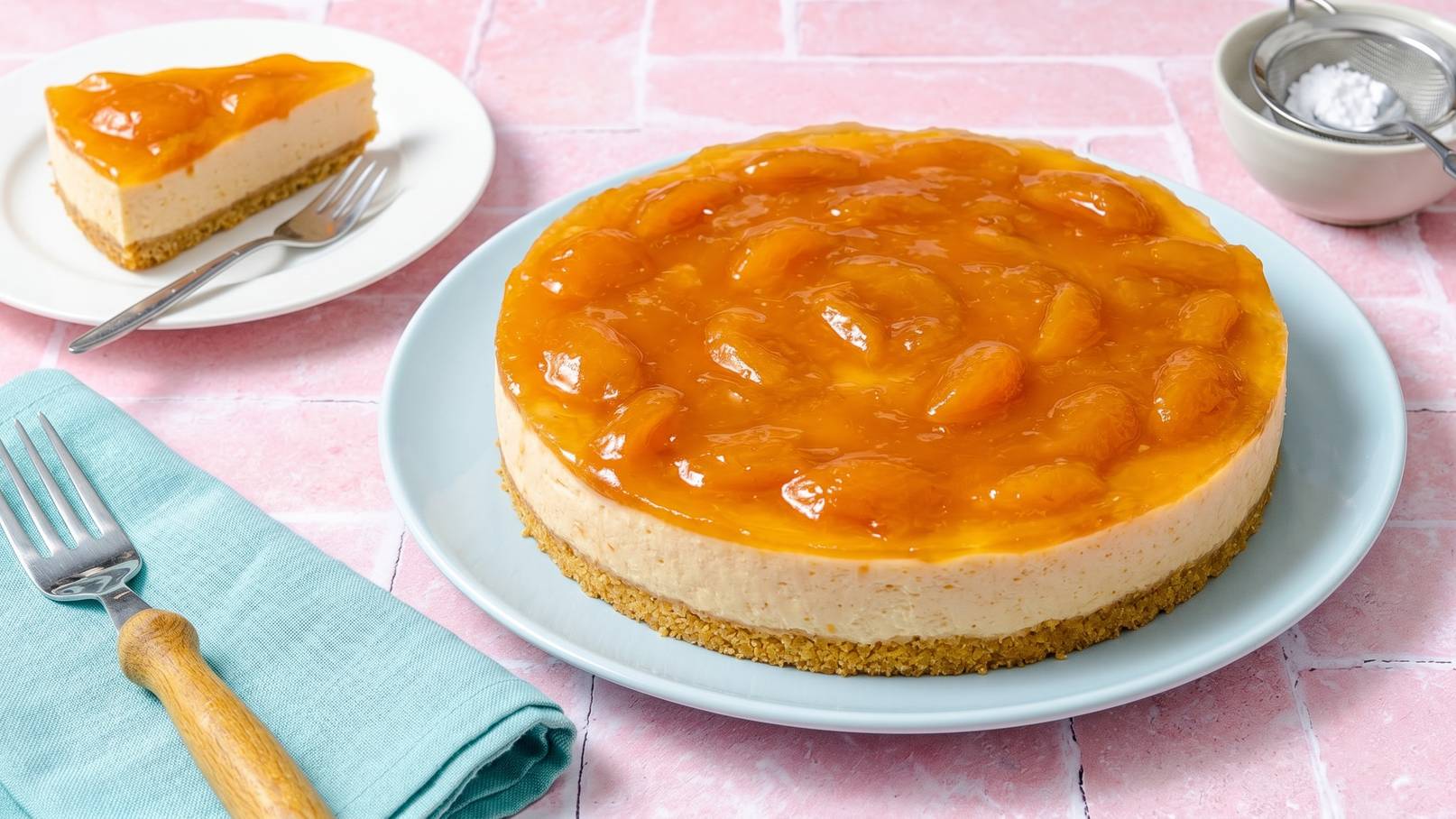 Auf einem hellen Teller liegt ein großer Käsekuchen mit einer glänzenden Schicht Aprikosenüberzug. Daneben steht ein kleineres Stück Kuchen auf einem weißen Teller. Eine Gabel und ein mintgrünes Servietten sind ebenfalls sichtbar. Der Hintergrund ist in sanften Rosa-Tönen.