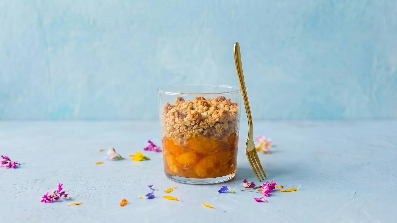 Ein Glas mit Schichtdessert: unten fruchtige Füllung, darüber knuspriges Müsli. Eine goldene Gabel steht neben dem Glas. Bunte Blütenblätter liegen dekorativ im Hintergrund. Der Hintergrund ist in sanften Blau- und Pastelltönen gehalten.