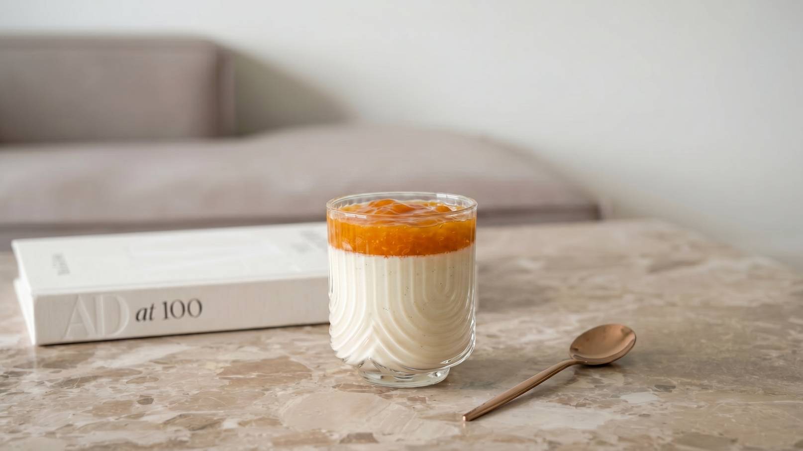 Ein Glas Dessert mit einer cremigen weißen Schicht und einer leuchtend orangefarbenen Fruchtsoße obenauf steht auf einem marmorierten Tisch. Neben dem Glas liegt ein kleiner Löffel. Im Hintergrund ist ein unscharfes, helles Sofa zu erkennen.